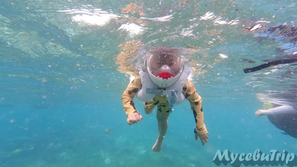 A happy child swimming in the clear blue waters of Hilutungan Marine Sanctuary wearing a shark mask.